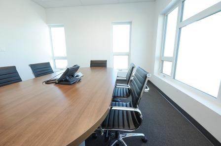 A close up of the boardroom, can see a phone sitting on a round wooden table, 5 black chairs tucked in and three bright windows. 